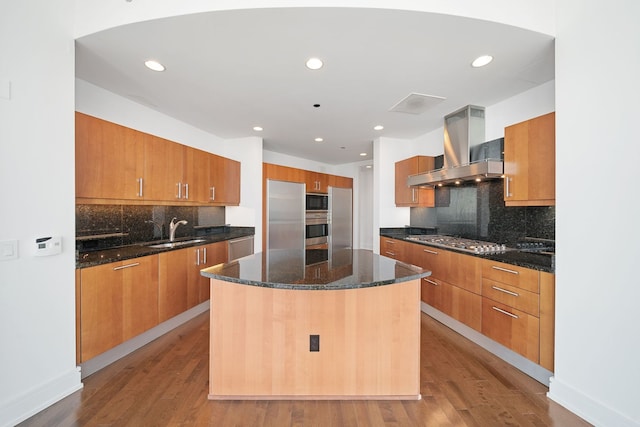 kitchen with a sink, wall chimney exhaust hood, brown cabinetry, backsplash, and appliances with stainless steel finishes