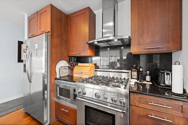 kitchen with wall chimney range hood, hardwood / wood-style floors, stainless steel appliances, tasteful backsplash, and dark stone counters