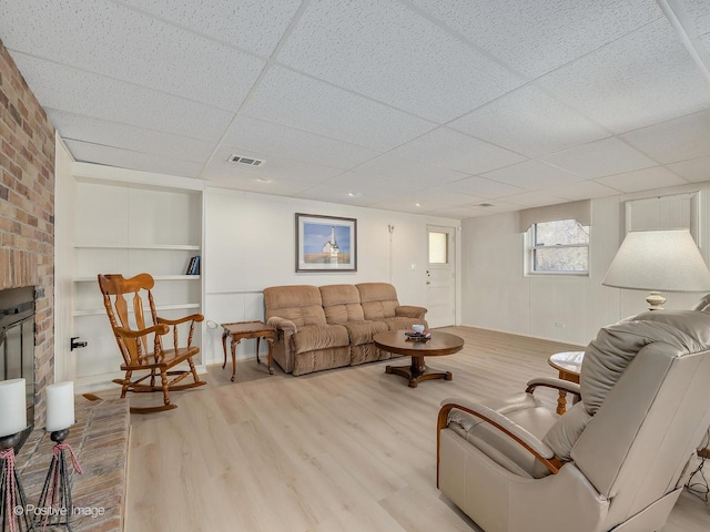 living room with a brick fireplace, a drop ceiling, and light wood-type flooring