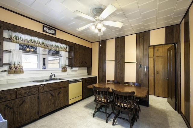 kitchen with dark brown cabinetry, sink, wooden walls, dishwashing machine, and ceiling fan