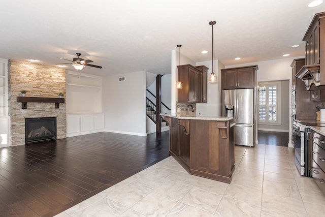 kitchen with a stone fireplace, dark brown cabinetry, stainless steel appliances, a peninsula, and visible vents