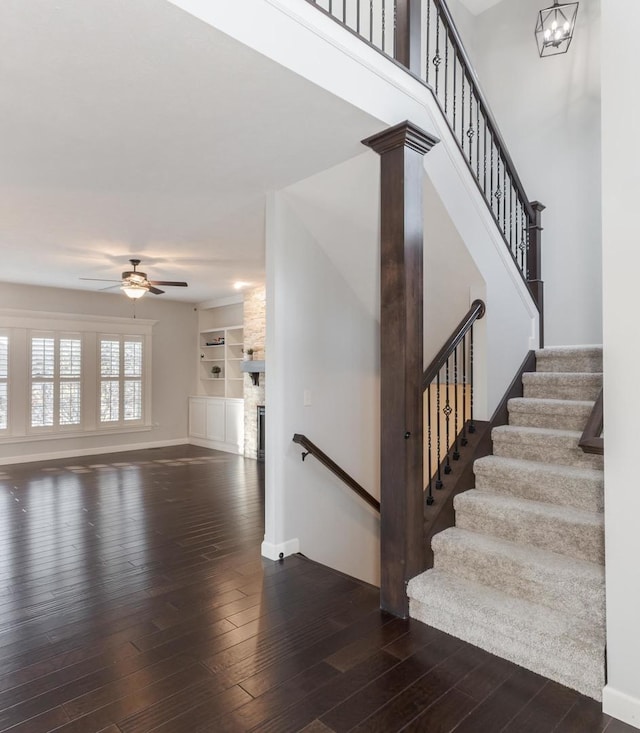 staircase with built in features, hardwood / wood-style flooring, a stone fireplace, baseboards, and ceiling fan with notable chandelier