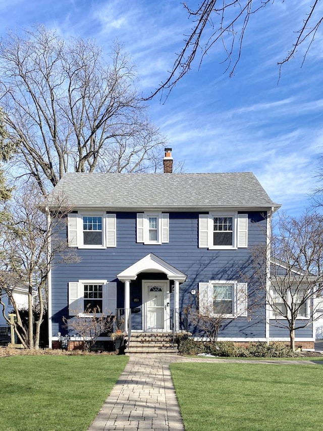 colonial house featuring roof with shingles, a front lawn, and a chimney