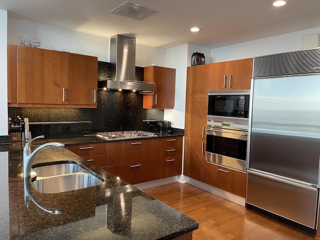 kitchen featuring sink, island range hood, built in appliances, light wood-type flooring, and dark stone counters