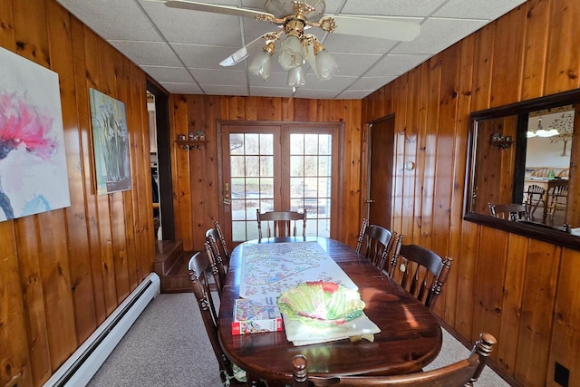 carpeted dining area with ceiling fan, a baseboard radiator, a drop ceiling, and wood walls