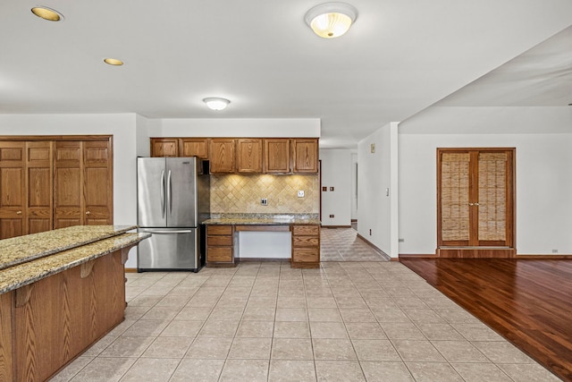 kitchen featuring tasteful backsplash, brown cabinetry, freestanding refrigerator, light tile patterned flooring, and light stone countertops