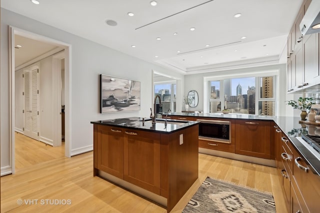 kitchen featuring sink, black microwave, a center island with sink, a raised ceiling, and light hardwood / wood-style floors