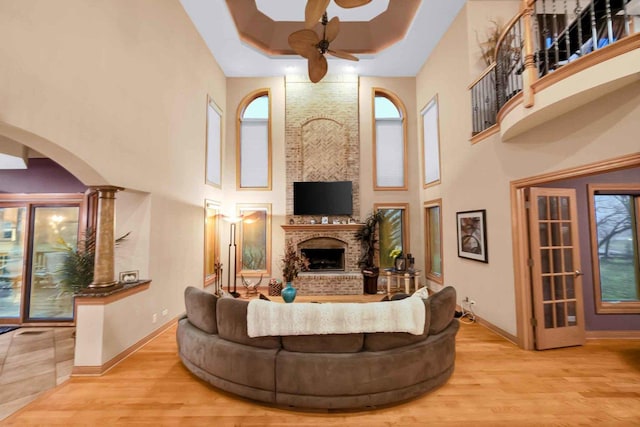 living room featuring ceiling fan, decorative columns, a brick fireplace, a raised ceiling, and light wood-type flooring