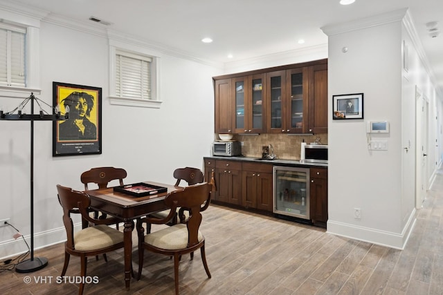 bar featuring tasteful backsplash, wine cooler, ornamental molding, dark brown cabinets, and light hardwood / wood-style flooring