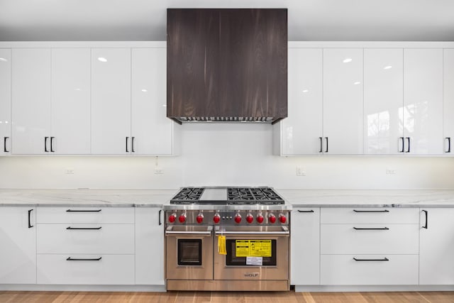 kitchen featuring white cabinetry, double oven range, wall chimney range hood, and light stone countertops