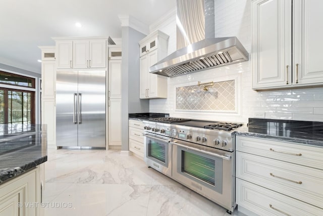 kitchen featuring dark stone countertops, white cabinetry, premium appliances, ornamental molding, and wall chimney exhaust hood