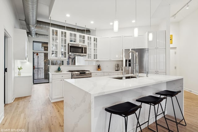 kitchen with pendant lighting, stainless steel appliances, light stone counters, an island with sink, and white cabinets