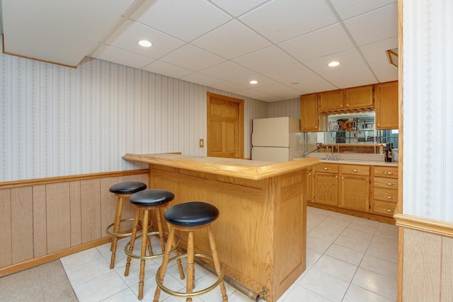 kitchen featuring light tile patterned flooring, a kitchen bar, a paneled ceiling, white refrigerator, and kitchen peninsula