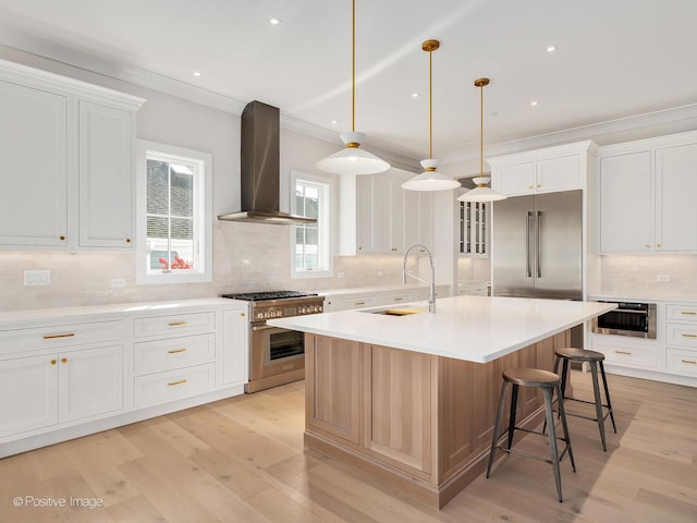 kitchen with high end appliances, pendant lighting, wall chimney range hood, a kitchen island with sink, and white cabinets