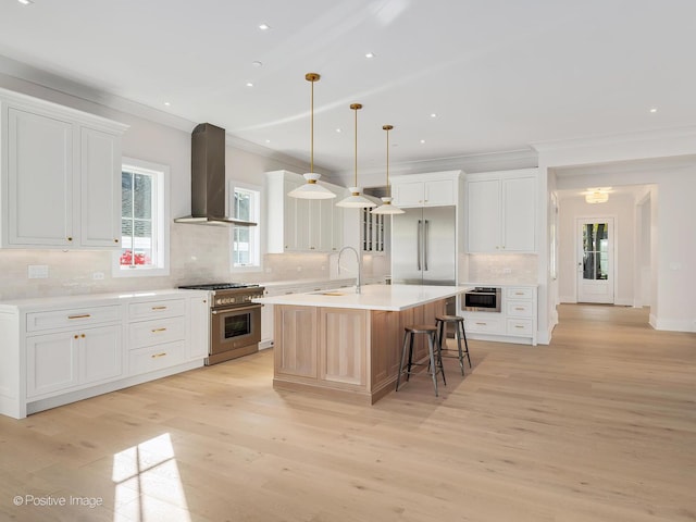 kitchen featuring wall chimney exhaust hood, white cabinetry, premium appliances, pendant lighting, and a kitchen island with sink