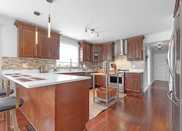 kitchen featuring wall chimney range hood, hanging light fixtures, stainless steel appliances, dark hardwood / wood-style flooring, and kitchen peninsula