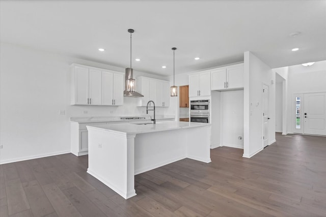 kitchen featuring white cabinets, stainless steel double oven, hanging light fixtures, a center island with sink, and wall chimney range hood