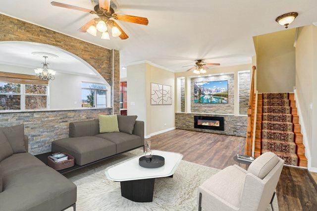 living area featuring ornamental molding, ceiling fan with notable chandelier, wood finished floors, a glass covered fireplace, and baseboards