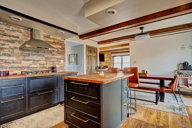 kitchen featuring butcher block countertops, wall chimney range hood, a breakfast bar area, stainless steel gas cooktop, and light wood-type flooring