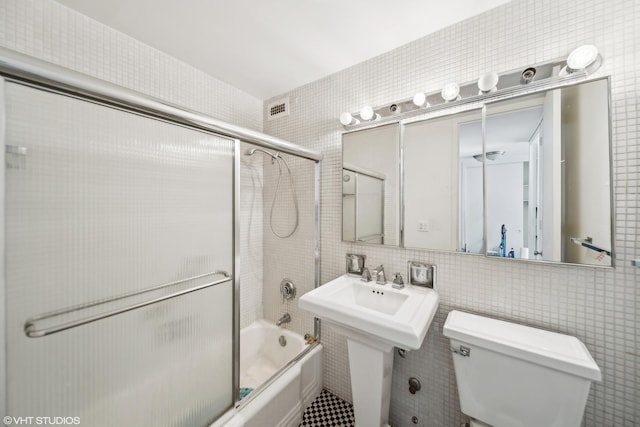 bathroom featuring toilet, combined bath / shower with glass door, and tile walls