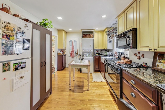 kitchen featuring stacked washer and dryer, light wood-type flooring, dark stone counters, and black appliances