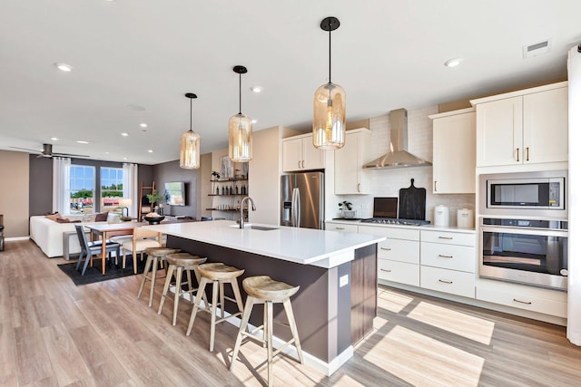 kitchen with an island with sink, sink, hanging light fixtures, stainless steel appliances, and wall chimney range hood