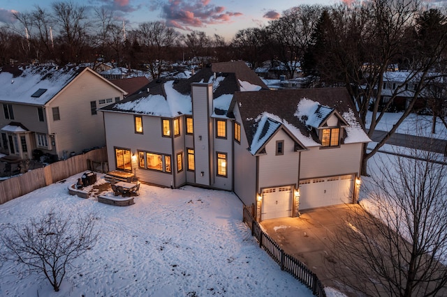 snow covered house featuring a chimney, an outdoor fire pit, fence, a garage, and driveway