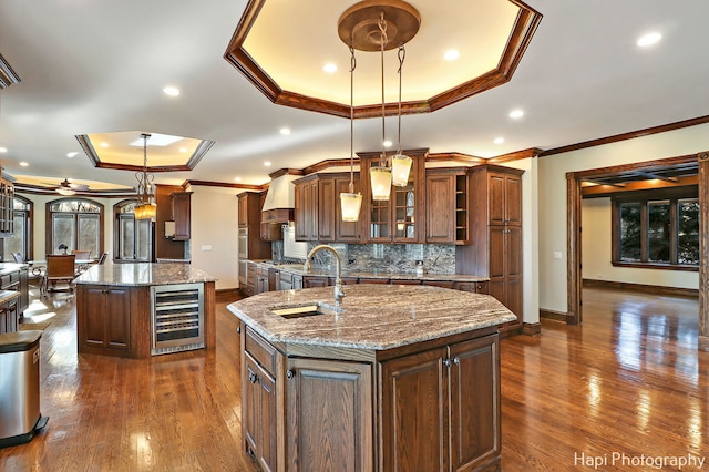 kitchen featuring wine cooler, sink, a center island with sink, a tray ceiling, and pendant lighting