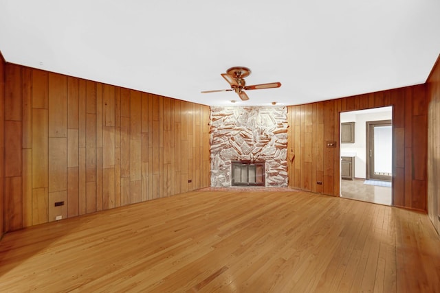 unfurnished living room with a stone fireplace, wood walls, ceiling fan, and light wood-type flooring