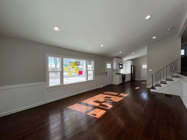 unfurnished living room with stairs, vaulted ceiling, dark wood finished floors, and a wainscoted wall