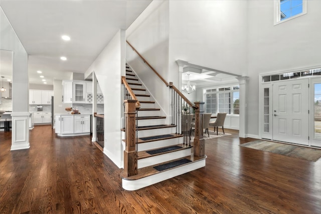 foyer entrance with dark wood-type flooring, a towering ceiling, decorative columns, and a notable chandelier