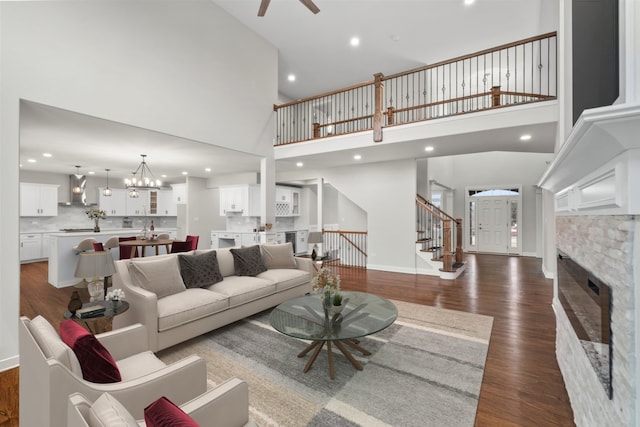 living room featuring a stone fireplace, dark wood-type flooring, a towering ceiling, and ceiling fan with notable chandelier