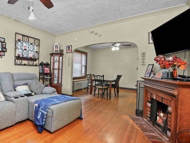 living room featuring hardwood / wood-style floors, ceiling fan, crown molding, a brick fireplace, and a textured ceiling