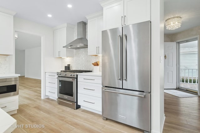 kitchen featuring appliances with stainless steel finishes, white cabinetry, decorative backsplash, light wood-type flooring, and wall chimney exhaust hood