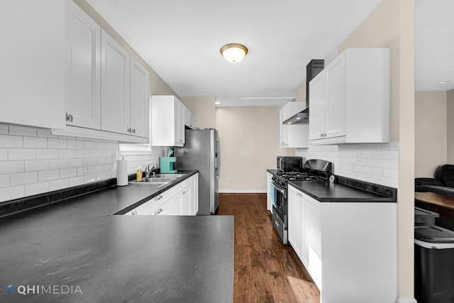 kitchen featuring dark countertops, appliances with stainless steel finishes, white cabinetry, a sink, and wall chimney exhaust hood