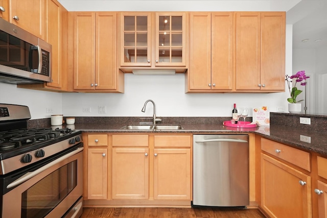 kitchen with stainless steel appliances, glass insert cabinets, a sink, dark stone countertops, and light wood-type flooring