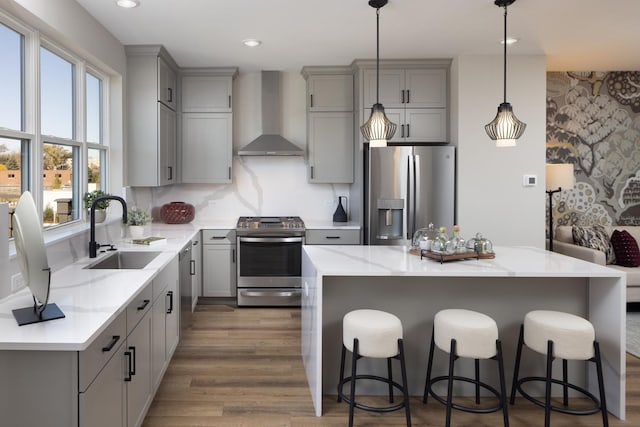 kitchen with wall chimney range hood, sink, appliances with stainless steel finishes, hanging light fixtures, and a kitchen island