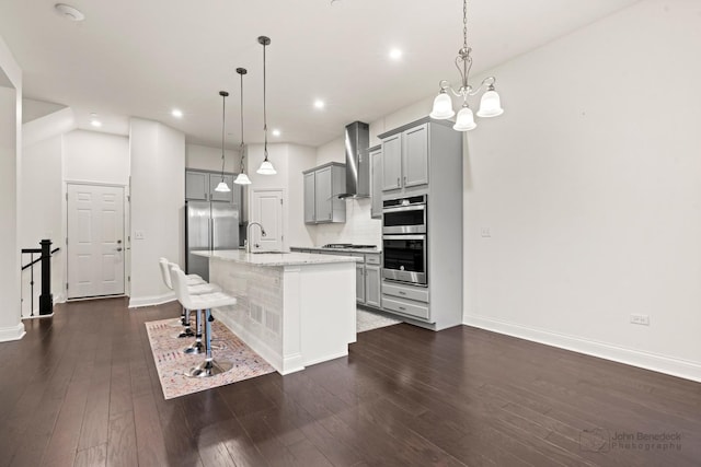 kitchen featuring gray cabinets, an island with sink, hanging light fixtures, and wall chimney range hood