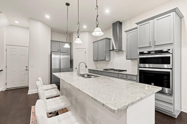 kitchen with appliances with stainless steel finishes, sink, hanging light fixtures, a center island with sink, and wall chimney exhaust hood