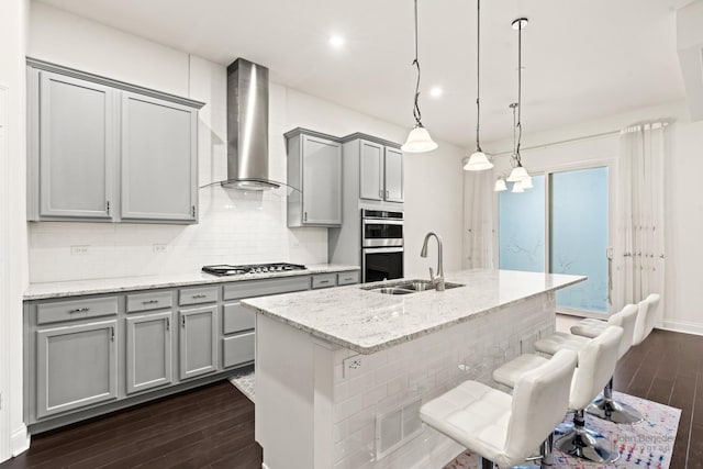 kitchen featuring wall chimney exhaust hood, a breakfast bar, sink, pendant lighting, and light stone countertops