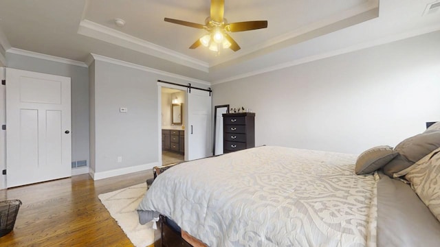 bedroom with connected bathroom, a barn door, a raised ceiling, hardwood / wood-style floors, and crown molding