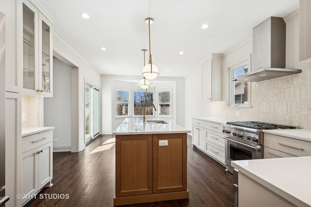 kitchen with sink, a center island with sink, hanging light fixtures, stainless steel stove, and wall chimney range hood