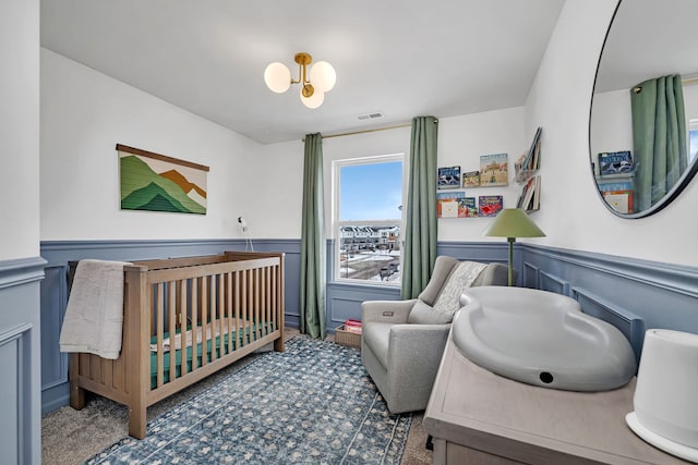 carpeted bedroom featuring a chandelier, a wainscoted wall, visible vents, and a nursery area