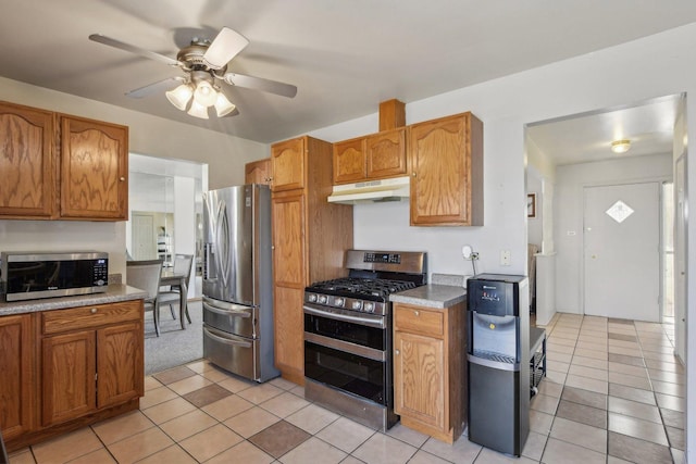 kitchen with under cabinet range hood, light tile patterned floors, brown cabinets, and appliances with stainless steel finishes