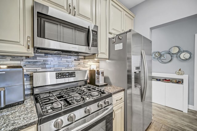 kitchen featuring appliances with stainless steel finishes, tasteful backsplash, light hardwood / wood-style floors, cream cabinetry, and dark stone counters