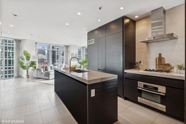 kitchen featuring sink, an island with sink, wall chimney range hood, oven, and backsplash