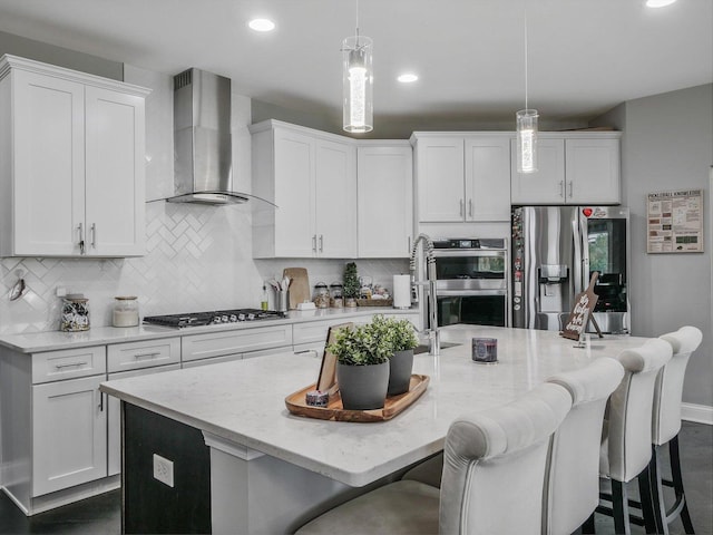 kitchen featuring appliances with stainless steel finishes, white cabinets, hanging light fixtures, a center island with sink, and wall chimney exhaust hood