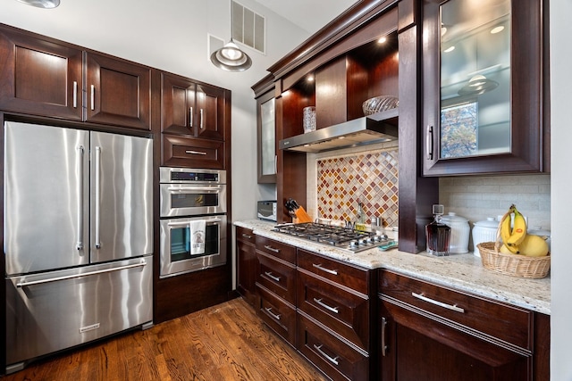 kitchen with dark wood-type flooring, appliances with stainless steel finishes, light stone counters, decorative backsplash, and wall chimney exhaust hood