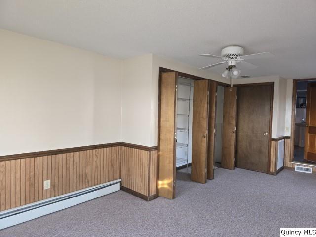 empty room with carpet floors, a baseboard heating unit, wooden walls, and ceiling fan