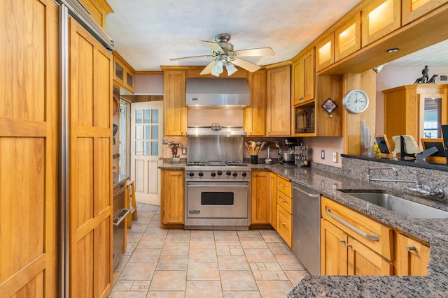 kitchen featuring sink, wall chimney range hood, ceiling fan, appliances with stainless steel finishes, and dark stone countertops